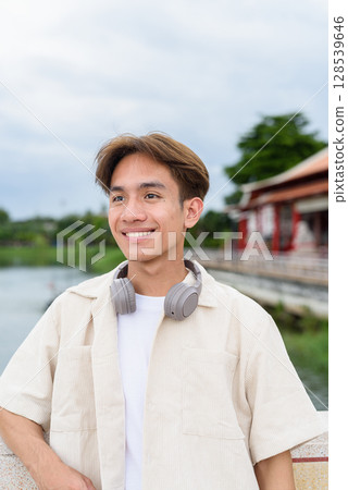 Asian non binary tourist person with headphones relaxing at park near lake 128539646