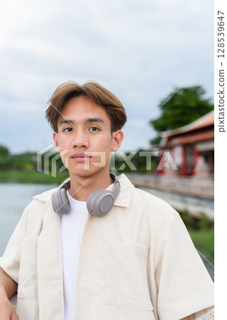 Asian non binary tourist person with headphones relaxing at park near lake 128539647