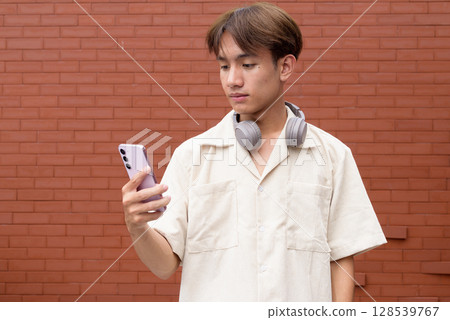 Non binary cute Asian young gay man using mobile phone against brick wall Non binary cute Asian young gay man using mobile phone against brick wall 128539767