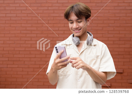 Non binary cute Asian young gay man using mobile phone against brick wall 128539768