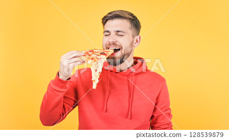 Happy Man In Red Hoodie Eating Pizza Slice On Vibrant Yellow Background For Fast Food And Lifestyle Visuals. Concept Of Casual Snacking, Comfort Food, Youth Culture, And Social Media Food Trends 128539879
