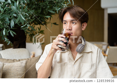 Non binary Asian student man sitting in modern coffee shop drinking cola 128539916