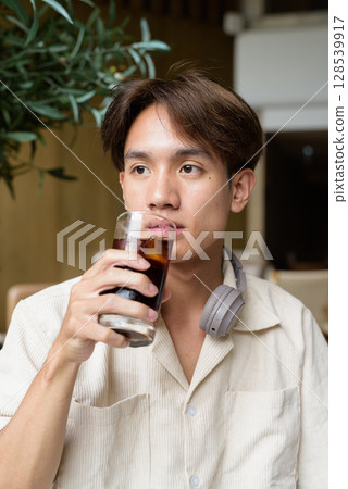 Non binary Asian student man sitting in modern coffee shop drinking cola 128539917