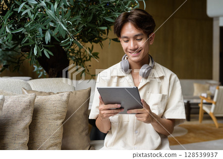 Non binary Asian student man sitting in modern coffee shop using digital tablet computer 128539937