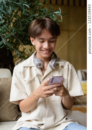 Non binary Asian student man sitting in modern coffee shop texting with phone 128539943