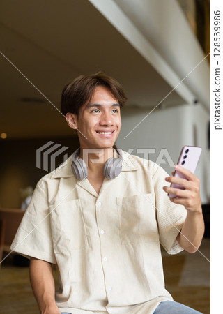 Non binary Asian student man sitting in modern coffee shop using phone 128539986