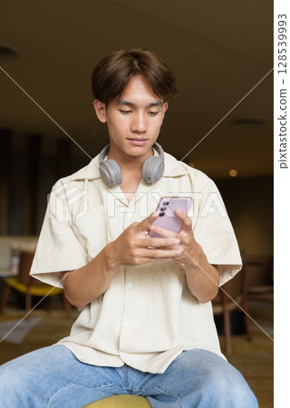 Non binary Asian student man sitting in modern coffee shop using phone 128539993