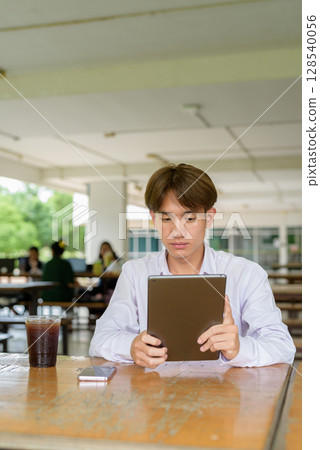 Non binary university student sitting in college campus using tablet computer Non binary university student sitting in college campus using tablet computer 128540056