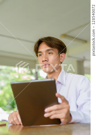 Non binary university student sitting in college campus using tablet computer Non binary university student sitting in college campus using tablet computer 128540072