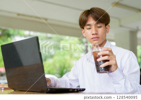 Non binary university student in college campus using laptop computer and drinking iced coffee Non binary university student in college campus using laptop computer and drinking iced coffee 128540095