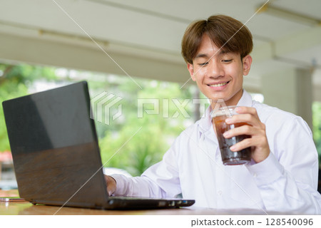 Non binary university student in college campus using laptop computer and drinking iced coffee Non binary university student in college campus using laptop computer and drinking iced coffee 128540096