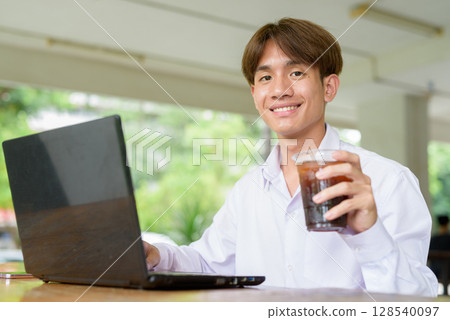 Non binary university student in college campus using laptop computer and drinking iced coffee 128540097
