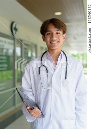 Young happy healthcare worker smiling and wearing lab coat in front of medical clinic Young happy healthcare worker smiling and wearing lab coat in front of medical clinic 128540182