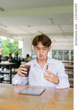 Non binary university student sitting in college campus using phone Non binary university student sitting in college campus using phone 128540269