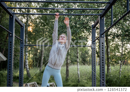 Young woman hanging on pull-up bar at outdoor gym in park Young woman hanging on pull-up bar at outdoor gym in park 128541372