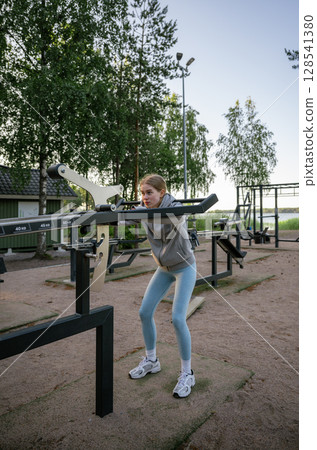 Young woman training on outdoor gym machine in park on a sunny day Young woman training on outdoor gym machine in park on a sunny day 128541380