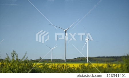 Wind turbines gracefully spinning in a bright blue sky over vibrant yellow fields at midday Wind turbines gracefully spinning in a bright blue sky over vibrant yellow fields at midday 128541401