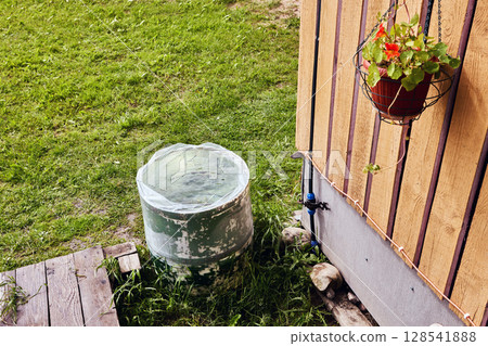 Rain barrel with cistern liner used for garden irrigation placed beside building below roof edge for harvested rain storage 128541888