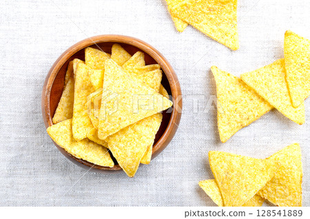 Tortilla chips in a wooden bowl on linen. Classic, crunchy snack food, made from corn tortillas, cut into triangle shaped wedges, fried in oil, and slightly salted. Close-up, from above, food photo. Tortilla chips in a wooden bowl on linen. Classic, crunchy snack food, made from corn tortillas, cut into triangle shaped wedges, fried in oil, and slightly salted. Close-up, from above, food photo. 128541889