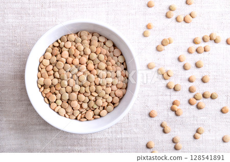 Plate lentils in a white bowl on linen fabric. Unshelled, dried and large green lentil variety, typically of flat shape, that hold their shape when cooked. Used in soups, stews, or salads. Food photo Plate lentils in a white bowl on linen fabric. Unshelled, dried and large green lentil variety, typically of flat shape, that hold their shape when cooked. Used in soups, stews, or salads. Food photo 128541891