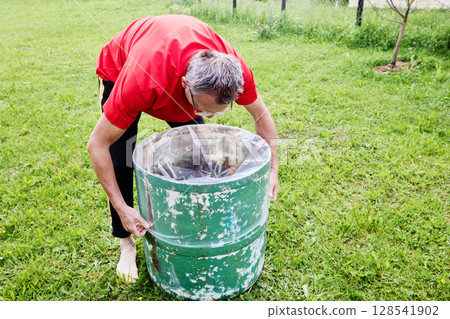 Pulling plastic liner tightly inside rain barrel, gardener ensures liner fits snugly along inner surface of watering barrel in backyard garden. Pulling plastic liner tightly inside rain barrel, gardener ensures liner fits snugly along inner surface of watering barrel in backyard garden. 128541902