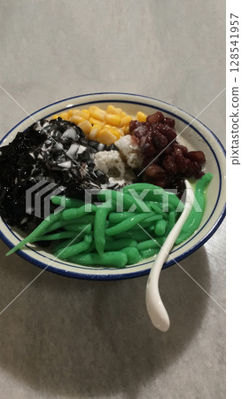 A bowl of cendol dessert with grass jelly, sweet corn and red bean topping. Cendol is a iced sweet dessert that is popular in southeast Asia.  128541957