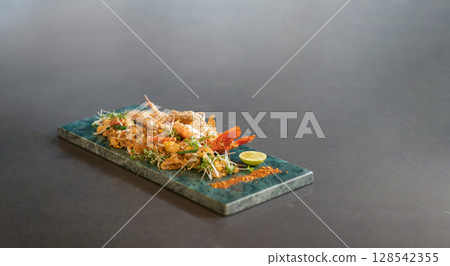 Lobster Pad Thai is served with yellow noodle, fried tofu, cress, bean sprouts, chilli power, garlic chives, and lime put on a rectangular plate. Lobster Pad Thai is served with yellow noodle, fried tofu, cress, bean sprouts, chilli power, garlic chives, and lime put on a rectangular plate. 128542355
