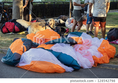 Paragliding group on grass with dog. Paragliders gather on grass with gear , golden retriever resting near colorful parachutes. Concept of paraglider community, shared passion, casual outdoor routine Paragliding group on grass with dog. Paragliders gather on grass with gear , golden retriever resting near colorful parachutes. Concept of paraglider community, shared passion, casual outdoor routine 128542798