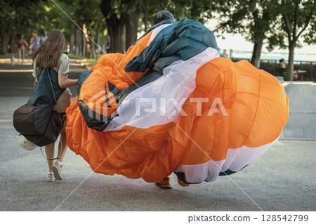 Paragliders carrying gear in park. Paragliders walk through park path with large wing folded in orange and white hues. Concept of post flight, sport lifestyle, shared adventure Paragliders carrying gear in park. Paragliders walk through park path with large wing folded in orange and white hues. Concept of post flight, sport lifestyle, shared adventure 128542799