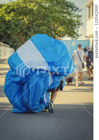 Man carrying parachute after flight. Paraglider carries large folded parachute on street after landing from flight. Concept of paraglider routine , sport lifestyle, real casual moment 128542800