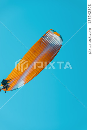 Orange paraglider in flight. Orange paraglider soars high in clear blue sky, capturing moment of tandem flight. Concept of paraglider adventure, sport hobby, freedom Orange paraglider in flight. Orange paraglider soars high in clear blue sky, capturing moment of tandem flight. Concept of paraglider adventure, sport hobby, freedom 128542808