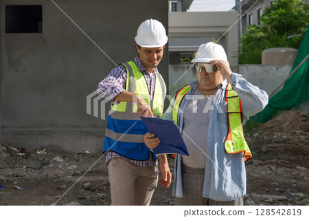 Two construction worker are reviewing project plan at a building site. One hold a clipboard while the other observe, with safety helmet and visibility vest enhancing their safety attire. 128542819