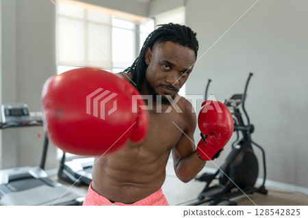 A muscular boxer wearing red gloves train hard in a gym, showcasing focus and determination as he prepare for his next match, surrounded by fitness equipment. A muscular boxer wearing red gloves train hard in a gym, showcasing focus and determination as he prepare for his next match, surrounded by fitness equipment. 128542825