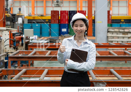 A dedicated female engineer dressed in safety gear stand smiling show finger thumb up, holding tablet computer amidst a sprawling factory. A dedicated female engineer dressed in safety gear stand smiling show finger thumb up, holding tablet computer amidst a sprawling factory. 128542826