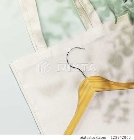 Sustainable shopping, buying or sales concept. Eco friendly spring still life with wooden hanger on white textile shopping bag on blue background with plant shadows. Flat lay 128542903