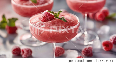 Refreshing raspberry frozen cocktail served in elegant glasses, garnished with mint leaves, surrounded by fresh raspberries and ice cubes 128543053