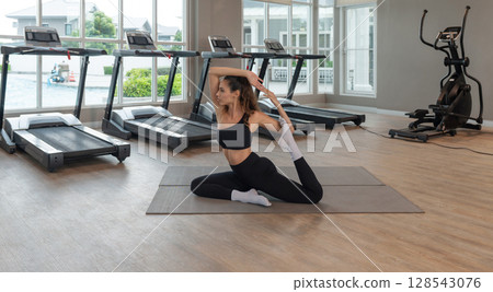 A woman perform King Pigeon Pose Variation (Mermaid Pose) on a mat in a contemporary gym. Sunlight stream through large window, highlighting her focused expression amidst fitness equipment. 128543076