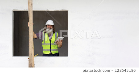 Senior engineer in hardhat and safety vest holding tablet computer stand in a window opening. Working atmosphere at construction site with scaffolding. 128543186