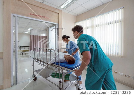 Two healthcare worker are hurriedly transferring a patient on a stretcher in a bright hospital emergency room during the day, illustrating urgency and teamwork in medical care. 128543226