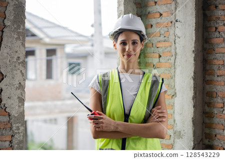 Female construction worker in hardhat and safety vest stand confidently with arms crossed in a partially built structure while holding a walkie-talkie during daytime. 128543229