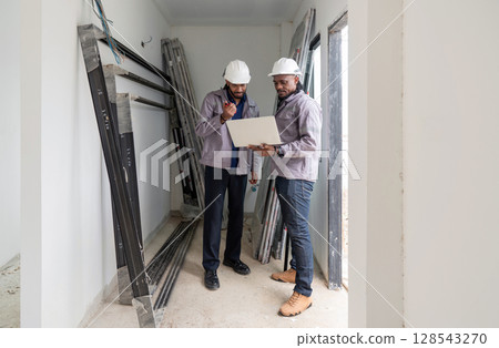 Two construction manager examine plan inside a developing project site. Various construction material surround them, indicating ongoing work. The early morning light create a focused atmosphere. 128543270