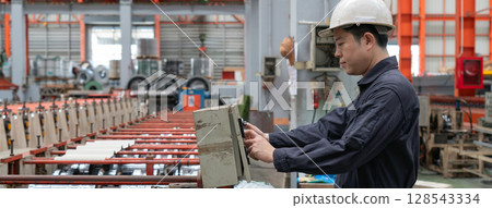 A man in a hardhat and mechanic coverall dust protection suit operates industrial equipment inside a busy factory setting, skillfully managing the machinery. 128543334