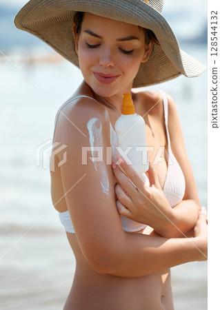 A beautiful woman smiles, applying sunscreen on skin. Skin and Body Care. Sun protection. Girl in bikini holding sun cream bottle on the beach. A female in a hat smears moisturizing lotion or suntan 128544132