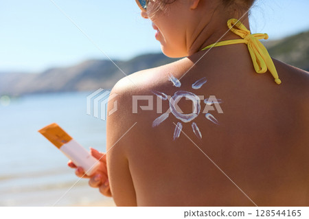 Skin care. Sun protection.  Woman With Suntan Lotion On Beach In Form Of The Sun. Portrait Of Female With  Drawn Sun On A Shoulder. Suncream. Girl Holding Moisturizing Sunblock and Apply Sun Cream 128544165