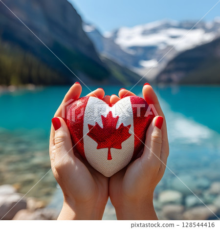 A person holds a heart-shaped object painted with the Canadian flag. The background features a serene lake and majestic mountains, symbolizing Canada Day. 128544416