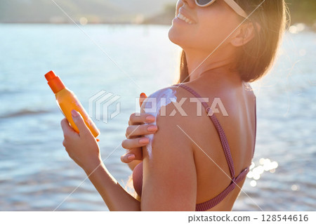 Beautiful Woman in Bikini Applying Sun Cream on Tanned  Shoulder. Sun Protection. Skin and Body Care. Girl Using Sunscreen to Skin. Portrait Of Female Holding Suntan Lotion or Moisturizing Sunblock. 128544616