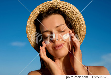 Female in hat applying  moisturizing lotion on skin. Beautiful Young woman with sun cream on face.Skin care. Sun protection. Suntan. 128544625