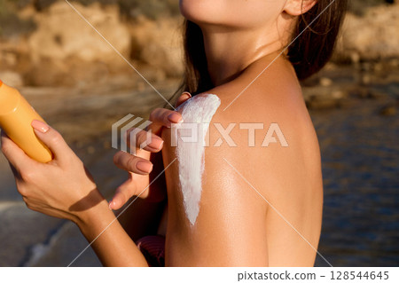 Sun cream on tanned shoulder. Sun protection. Beautiful woman in bikini applying  Solar Cream.  Skin and body care.  Portrait of female holding suntan lotion or moisturizing sunblock. 128544645
