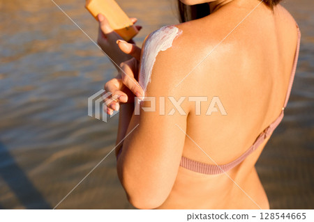 Young beautiful woman applying sunscreen on herself to protect her skin from the sun at the beach. 128544665