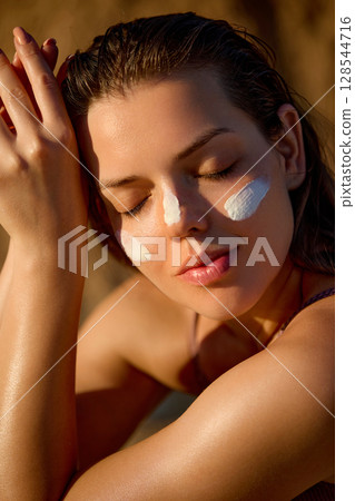 Suntan spf lotion. Happy lady applying sunscreen solar cream to her cheek, sitting on the beach. Sun cream protection. Skincare cosmetics model. Skin and Body Care Concept 128544716